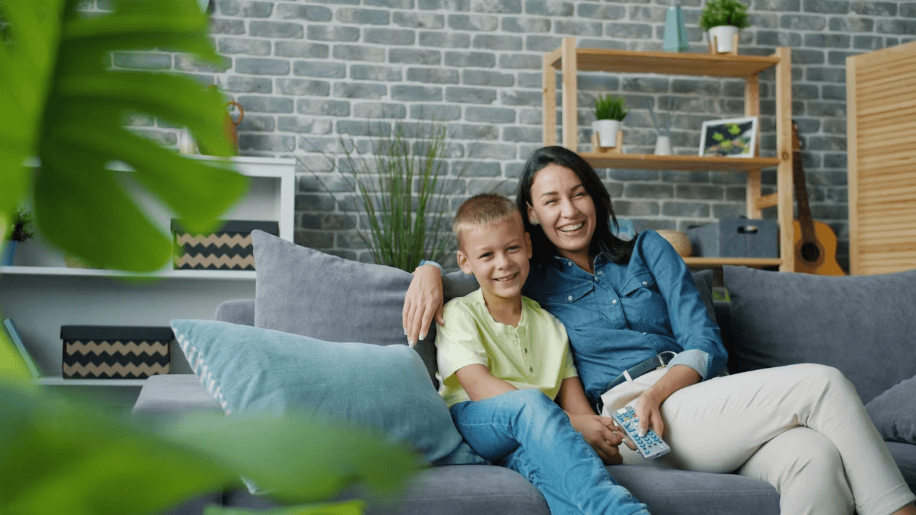 A mother and her son sitting together on a sofa, smiling while watching television